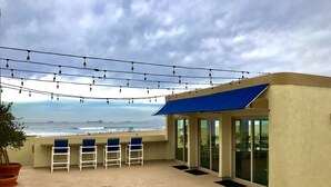 Terraço com vista para o oceano, com toldo azul, cadeiras brancas e luzes de corda.