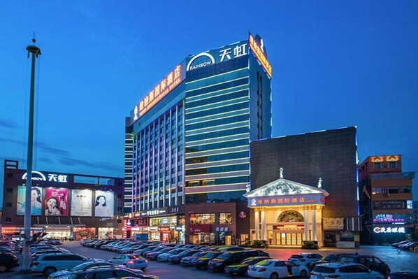 A multi-story hotel with a prominent 'Rainbow' sign, surrounded by a parking lot and various commercial establishments. - Venus International Hotel Shenzhen Gongming Square Metro Station Tianhong Branch (Shenzhen)