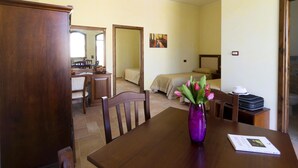 Una camera d'albergo con un tavolo da pranzo in legno, un vaso viola con fiori, un letto con copriletto color crema e un quadro appeso alla parete.