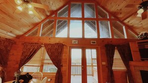 A cozy cabin interior with a high ceiling, wooden walls, a dining area with a table and chairs, a fireplace, and a large window with curtains. - Mountain Views ** Close2 Parkway/Gatlinburg/Dollywood ** | TreeHouse  (Sevierville)