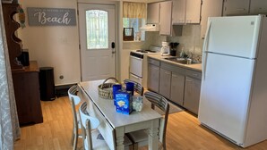A compact kitchen with a refrigerator, stove, and sink. There is a dining table with chairs and a basket. A sign on the wall reads "Life is better at the Beach." - Private beach; firepit, outdoor shower, AC, two kitchens, laundry, covered patio (Narragansett)