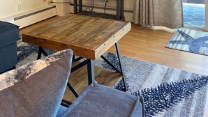 A living room with a wooden table, two grey armchairs, a TV on the wall, a bookshelf, and a window with curtains. - Private beach; firepit, outdoor shower, AC, two kitchens, laundry, covered patio (Narragansett)