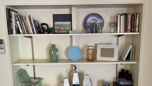 A bookshelf with books, a framed picture, decorative jars, and a small chair. - Private beach; firepit, outdoor shower, AC, two kitchens, laundry, covered patio (Narragansett)