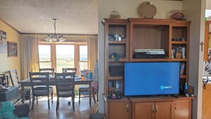 Ein Wohnzimmer mit Fernseher, Holzschränken, einem Essbereich mit Tisch und Stühlen sowie Blick ins Freie durch große Fenster.