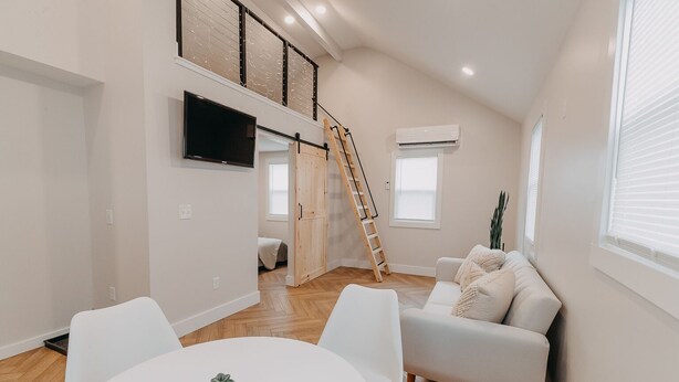 A spacious living room with a high ceiling, a white couch, a dining table with a small plant, and a wooden ladder leading to a loft area. - The Loft w/outdoor fire pit (Grandville)