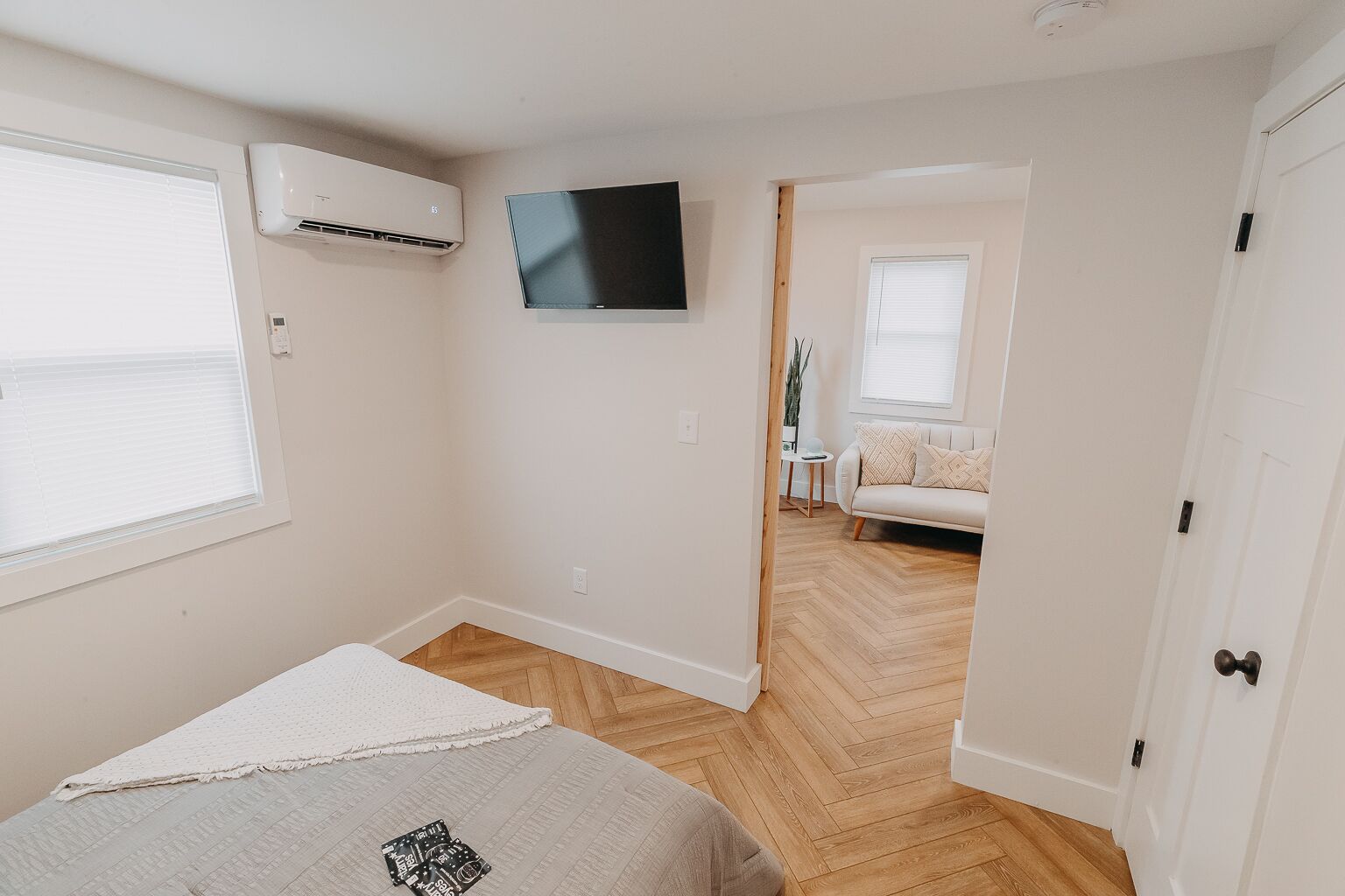 Un dormitorio con cama, aire acondicionado, televisión y vista a otra habitación con sofá.