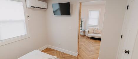 Un dormitorio con cama, aire acondicionado, televisión y vista a otra habitación con sofá.