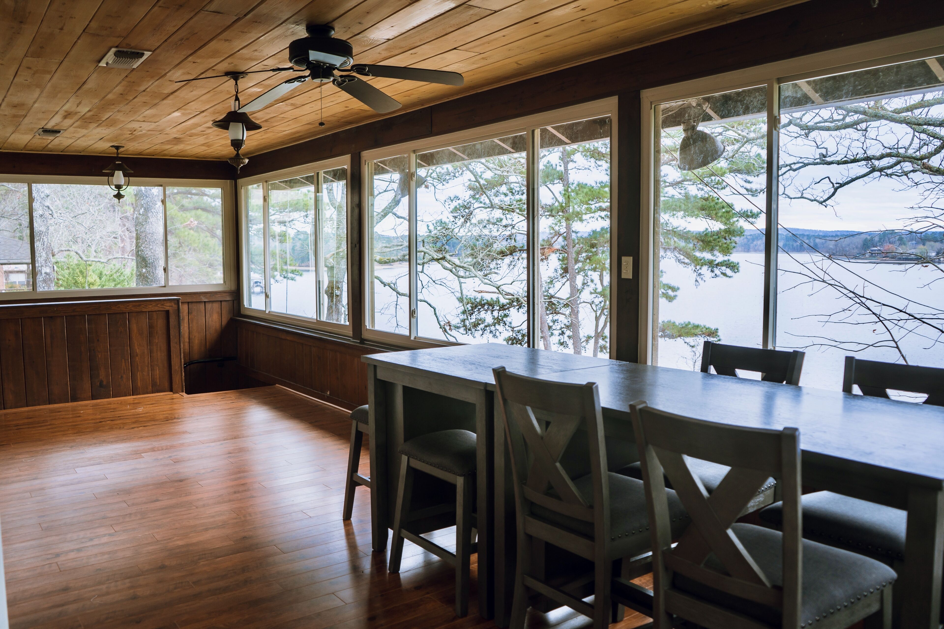 A dining area with a large table and chairs, wooden flooring, and large windows with a view of trees and a body of water.