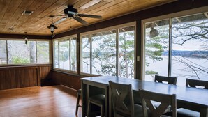 Un espace repas comprenant une grande table et des chaises, un plancher en bois et de grandes fenêtres offrant une vue sur des arbres et une étendue d’eau.