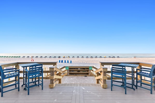 A wooden deck with blue chairs and tables, leading to a beach with a lifeguard tower and a clear blue sky.