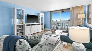 A modern living room with a flat-screen TV, a sofa, a coffee table, and a balcony with a view. - SunDestin Beach Resort Getaway With Pool, Hot Tub and Gulf Access (Destin)
