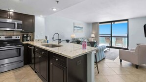 A modern kitchen with dark cabinets, stainless steel appliances, and a large island with a sink. There is a dining area with a sofa and chairs, and a large window offering a view.