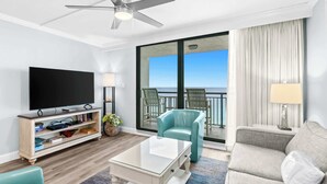 A modern hotel room with a flat-screen TV, a glass-top coffee table, a sofa, a balcony with a view, and a ceiling fan.