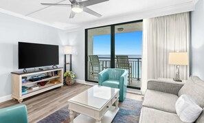 A modern hotel room with a flat-screen TV, a glass-top coffee table, a sofa, a balcony with a view, and a ceiling fan.