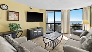 A living room with a sofa, a coffee table, a TV, a plant, and a view of the ocean.