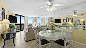 A modern living room with a glass dining table, white chairs, a sofa, and a ceiling fan.