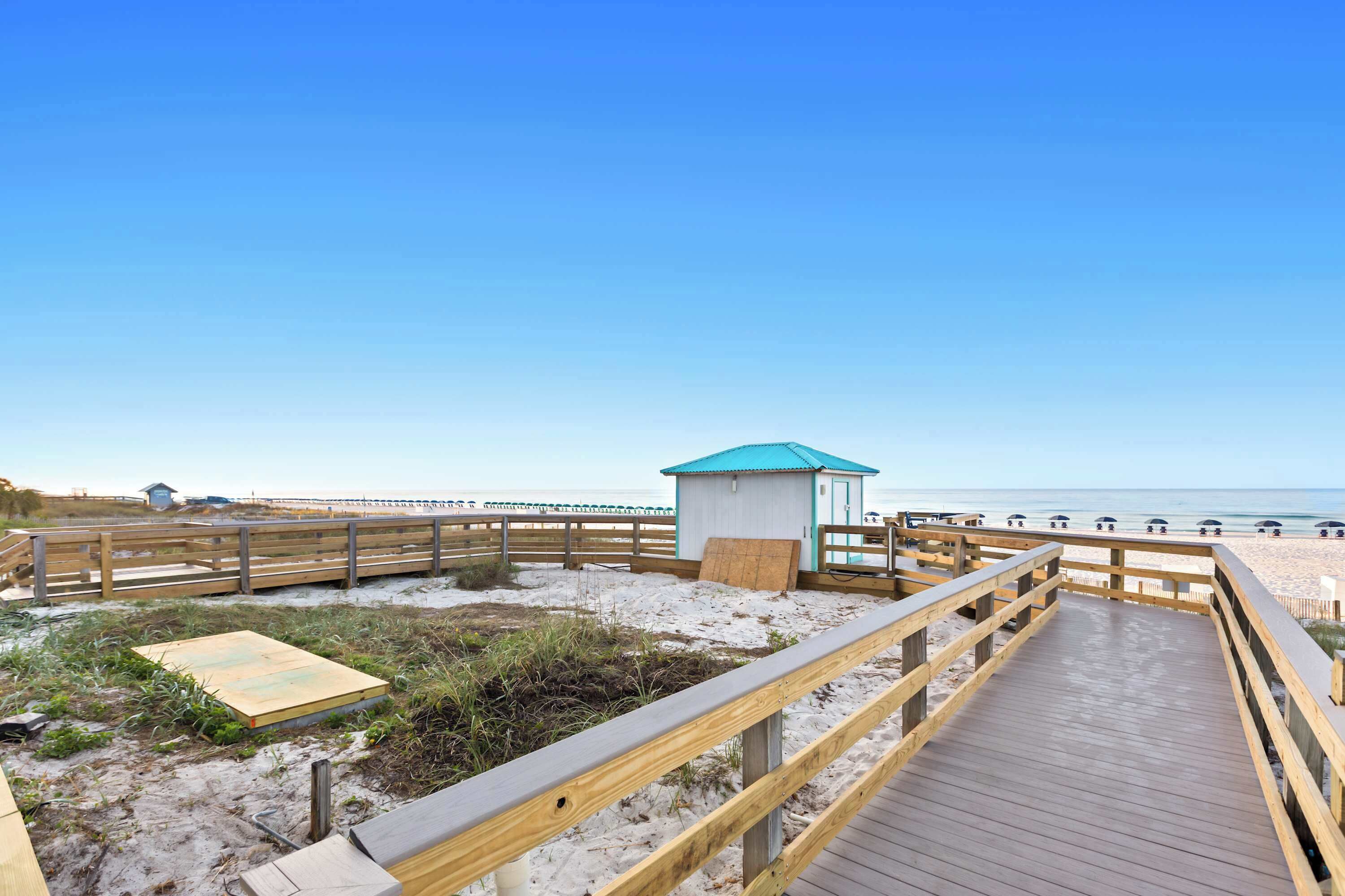 Een houten looppad dat naar een strand leidt, met een klein blauw gebouw, mogelijk een duikerspost.