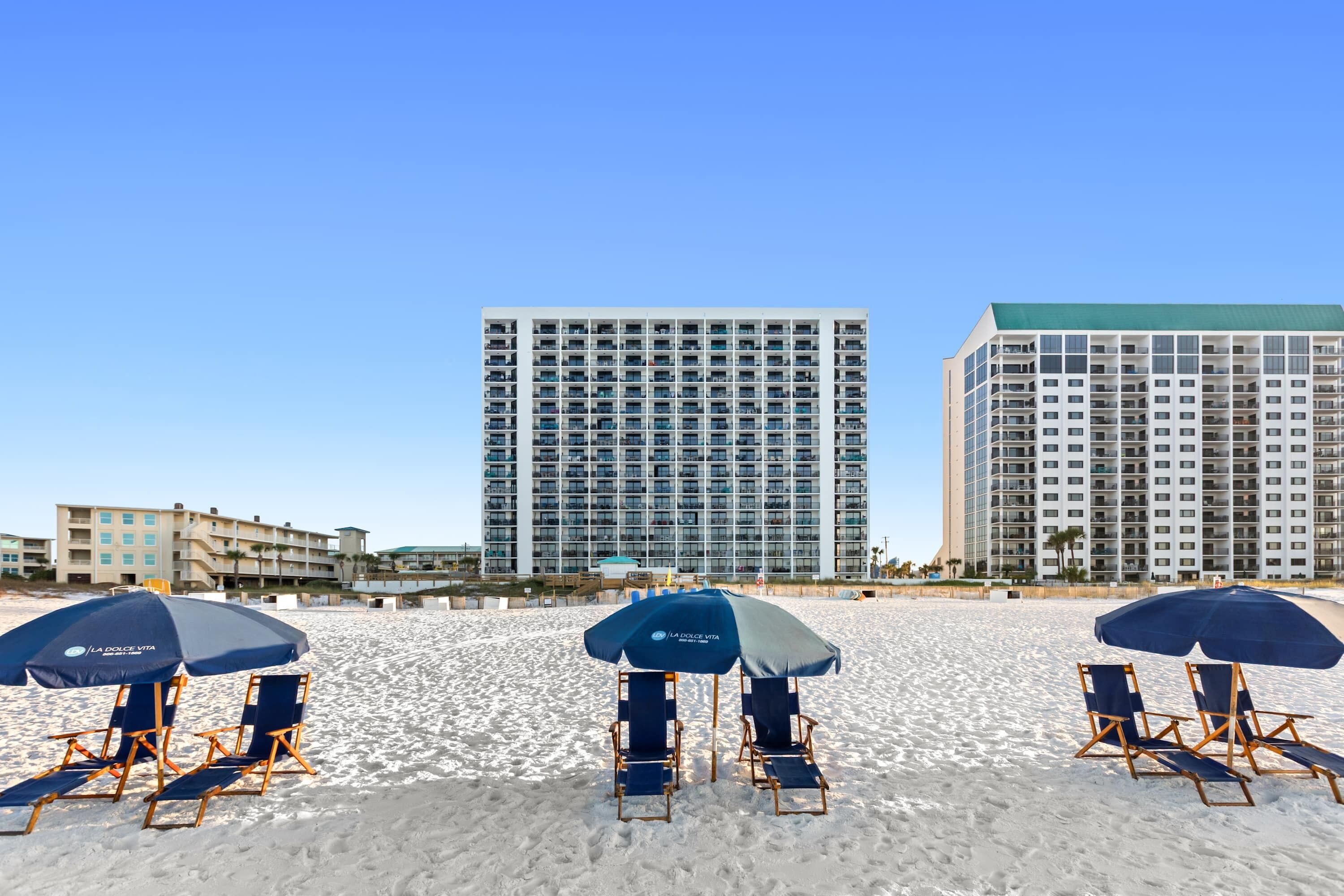 A beach with three blue umbrellas and beach chairs, a tall building in the background, and a clear blue sky.