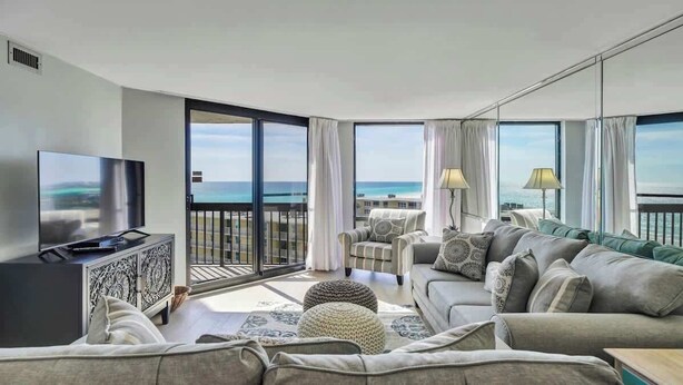 A modern living room with a large flat-screen TV, a sofa set, and a view of the ocean through large windows.