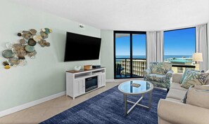 A modern living room with a flat-screen TV, a glass coffee table, a sofa, and a balcony with a view of the ocean.