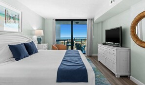 A hotel room with a bed, a television, a dresser, and a view of the ocean.