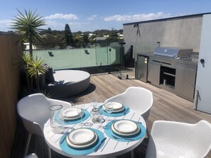 A round table set for four with white chairs, a grill, and a view of the surrounding area. - Beautiful new village apartment with roof deck- stone bath, 360  views & more (Barwon Heads)