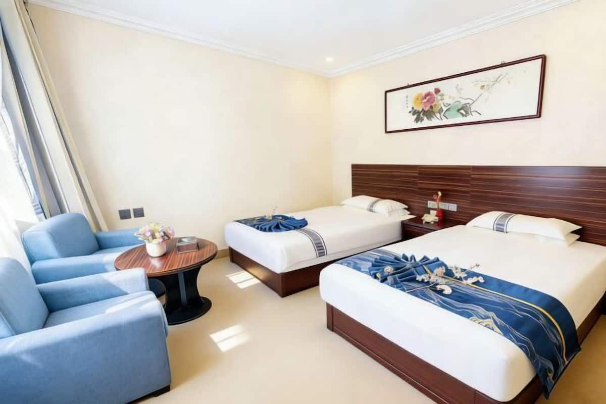 Habitación de hotel con dos camas, un sofá, una mesita con un florero y un cuadro en la pared.