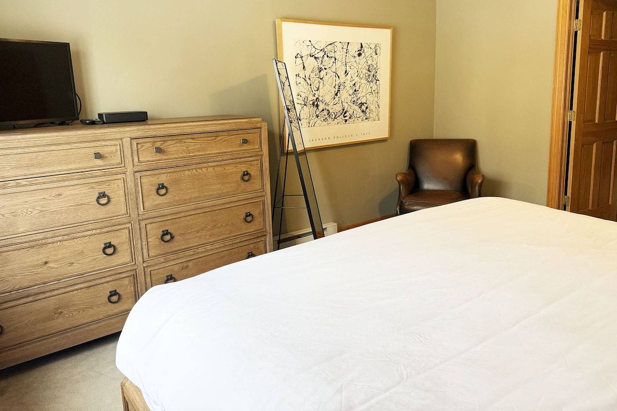 A bedroom with a wooden dresser, a bed with white bedding, a framed artwork on the wall, and a chair.
