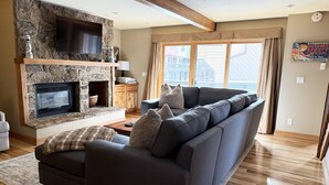 A modern living room with a stone fireplace, a large grey sofa, and a wooden floor. - 4 enSuites/2 King Beds-2 Car Garage-EZ Walk to Trails/Resort +Free Town Bus-W/D in Unit-Large Deck (Steamboat Springs)