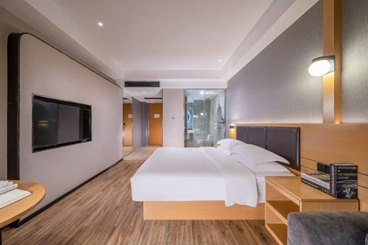 A modern hotel room with a large bed, a flat-screen TV mounted on the wall, a bedside table with a lamp, and a small seating area with a chair.