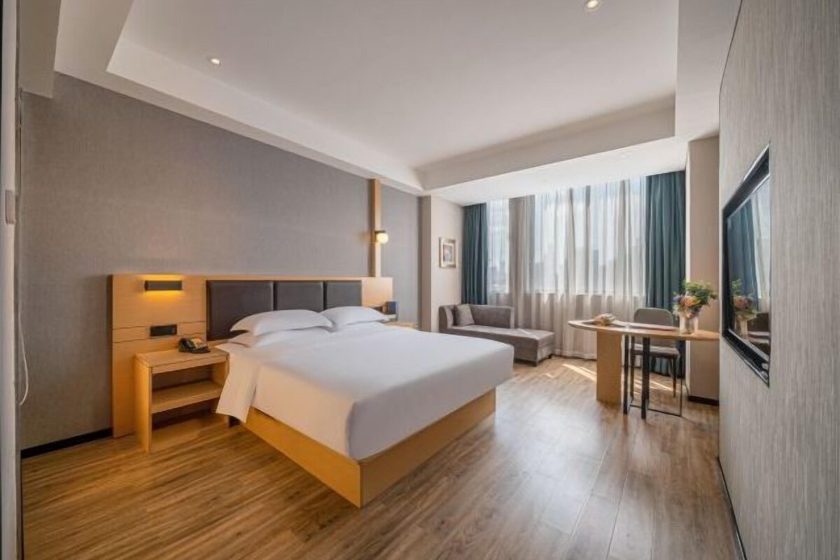 A modern hotel room with a large bed, bedside table, and a desk with a chair.