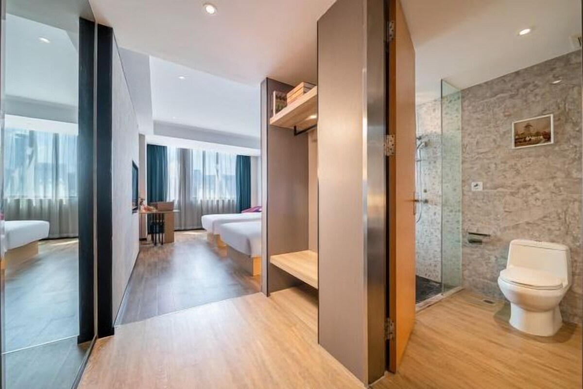 Una habitación de hotel moderna con suelo de madera, cabina de ducha de vidrio, inodoro blanco y cama con cabecera de madera.