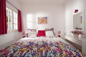 A bedroom with a floral bedspread, a painting on the wall, and a dresser with a vase of flowers.