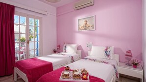 Et pink-farvet hotelværelse med to senge, et vindue med gardiner, en aircondition, et natbord med lampe og et bakke med te og blomster.