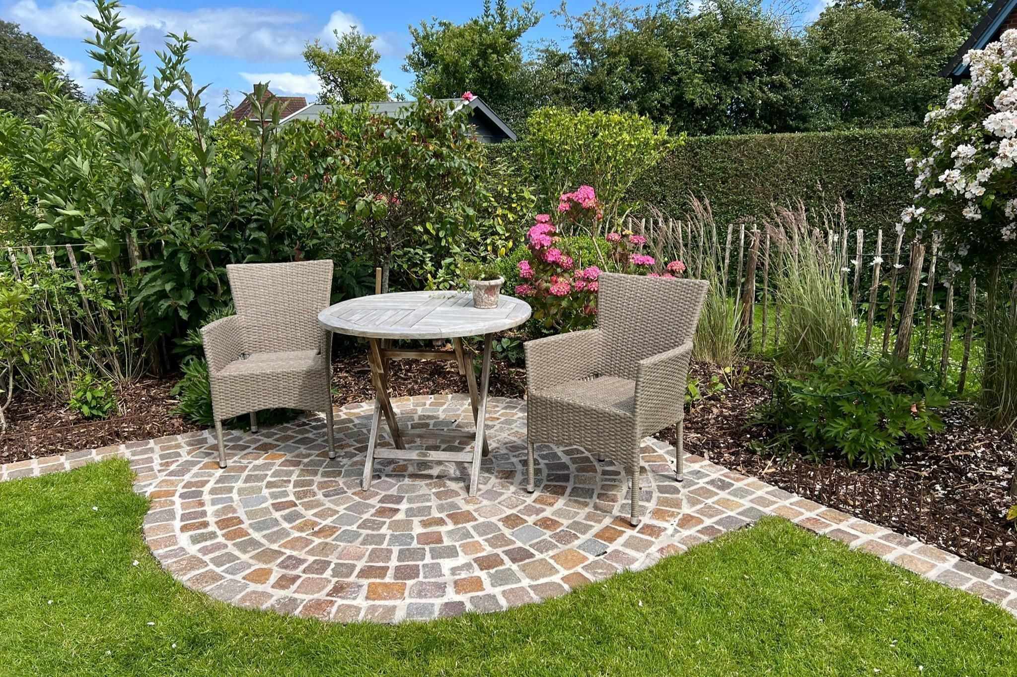 A patio with two wicker chairs and a table, surrounded by a mosaic-tiled floor and lush greenery.
