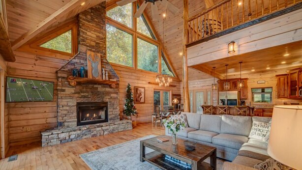 A spacious log cabin interior with a stone fireplace, large windows, a cozy living area with a sofa, and a kitchen with wooden cabinets. - Heaven on the Lake - Lakefront | Hot Tub | Fire Pit | Game Room (Blue Ridge)