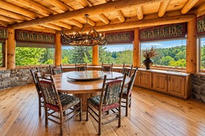 A wooden dining room with a round table and chairs, large windows with forest views, and a chandelier. - Lonesome Dove - Custom Western Style Log Lodge | Screened in outdoor living (Blue Ridge)