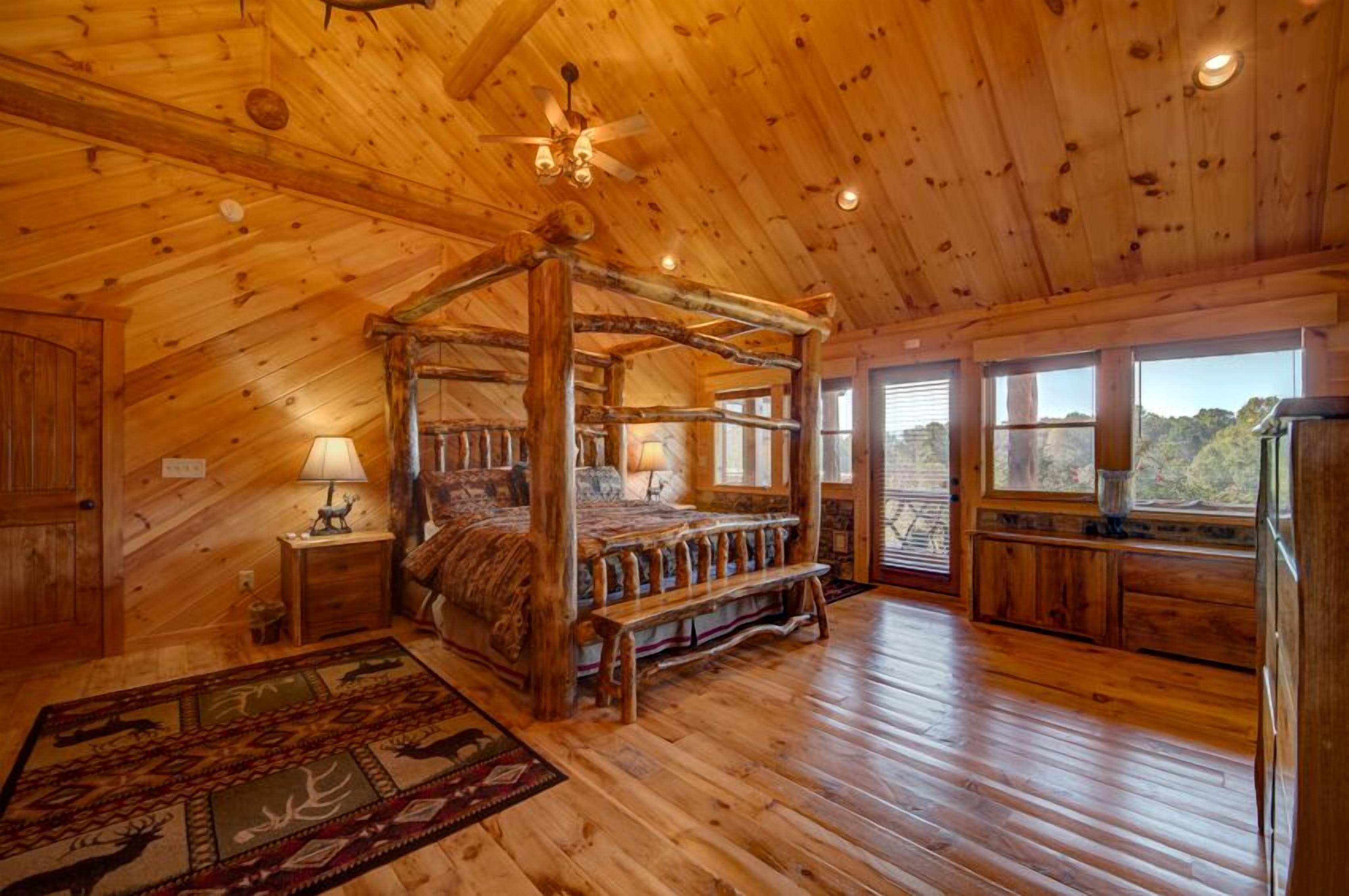 A rustic wooden cabin bedroom with a four-poster bed, a wooden ceiling, and large windows offering a view of the outdoors.