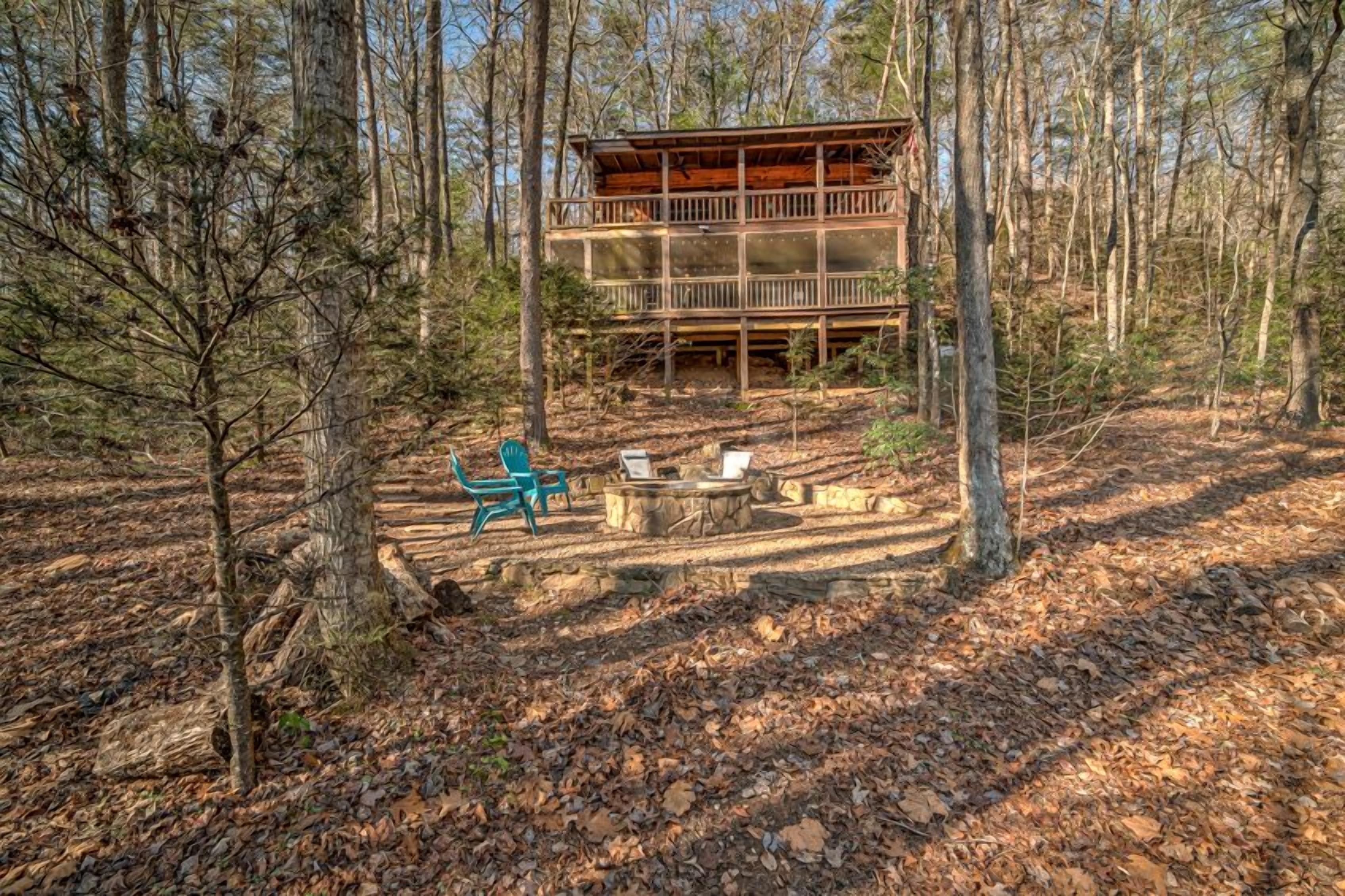 Une cabane dotée d’une terrasse, entourée d’arbres, et un foyer avec des chaises.