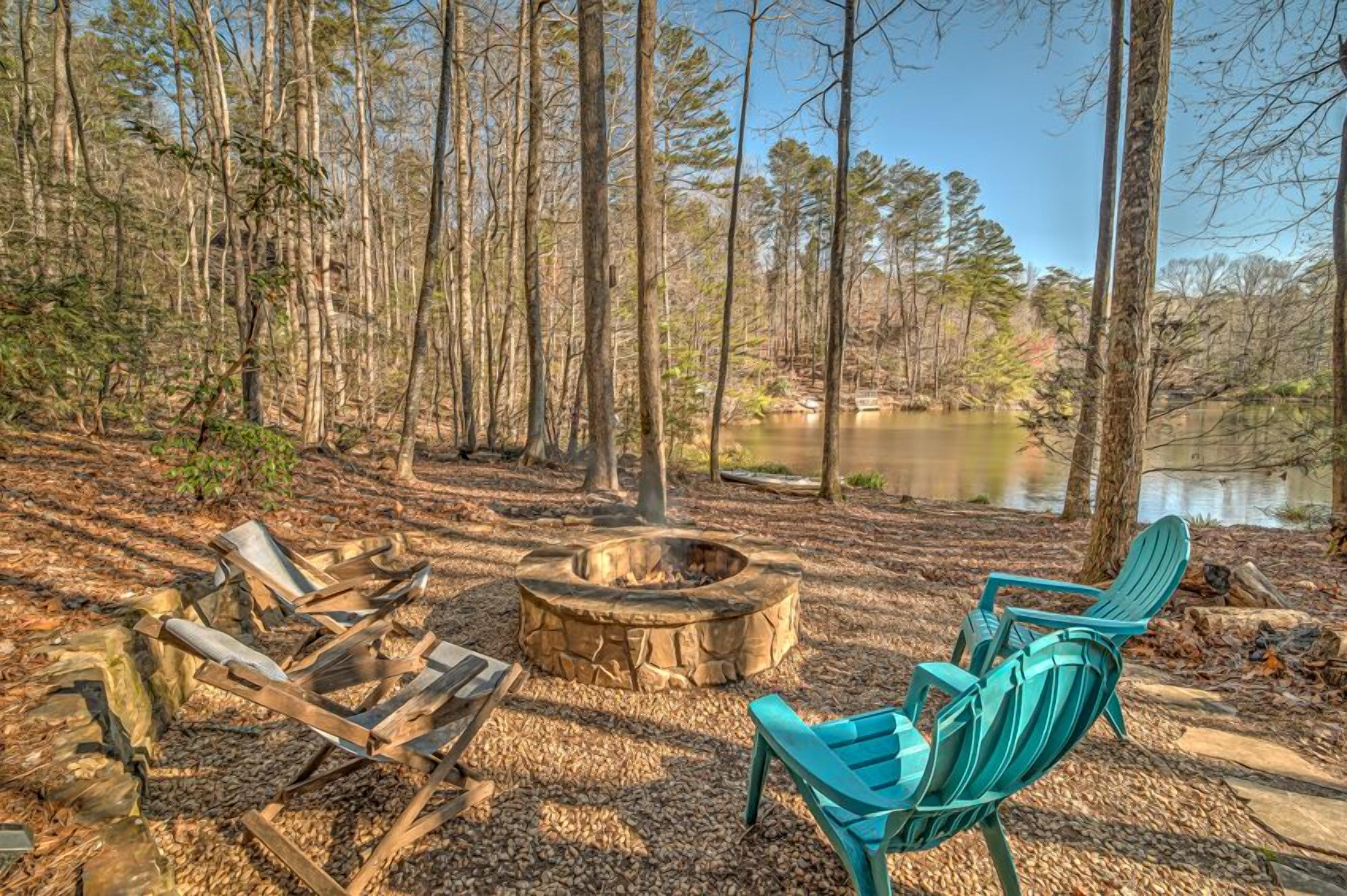 Um camping com lareira e duas cadeiras Adirondack perto de um lago rodeado por árvores.