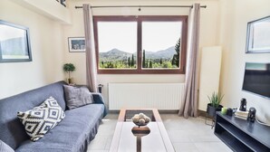 Uma sala de estar com um sofá, uma mesa de centro com objetos decorativos, uma TV em um móvel e uma janela com vista para montanhas.