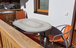 Een houten terras met een ronde tafel en oranje stoelen, een raam met zonwering, en een witte muur.