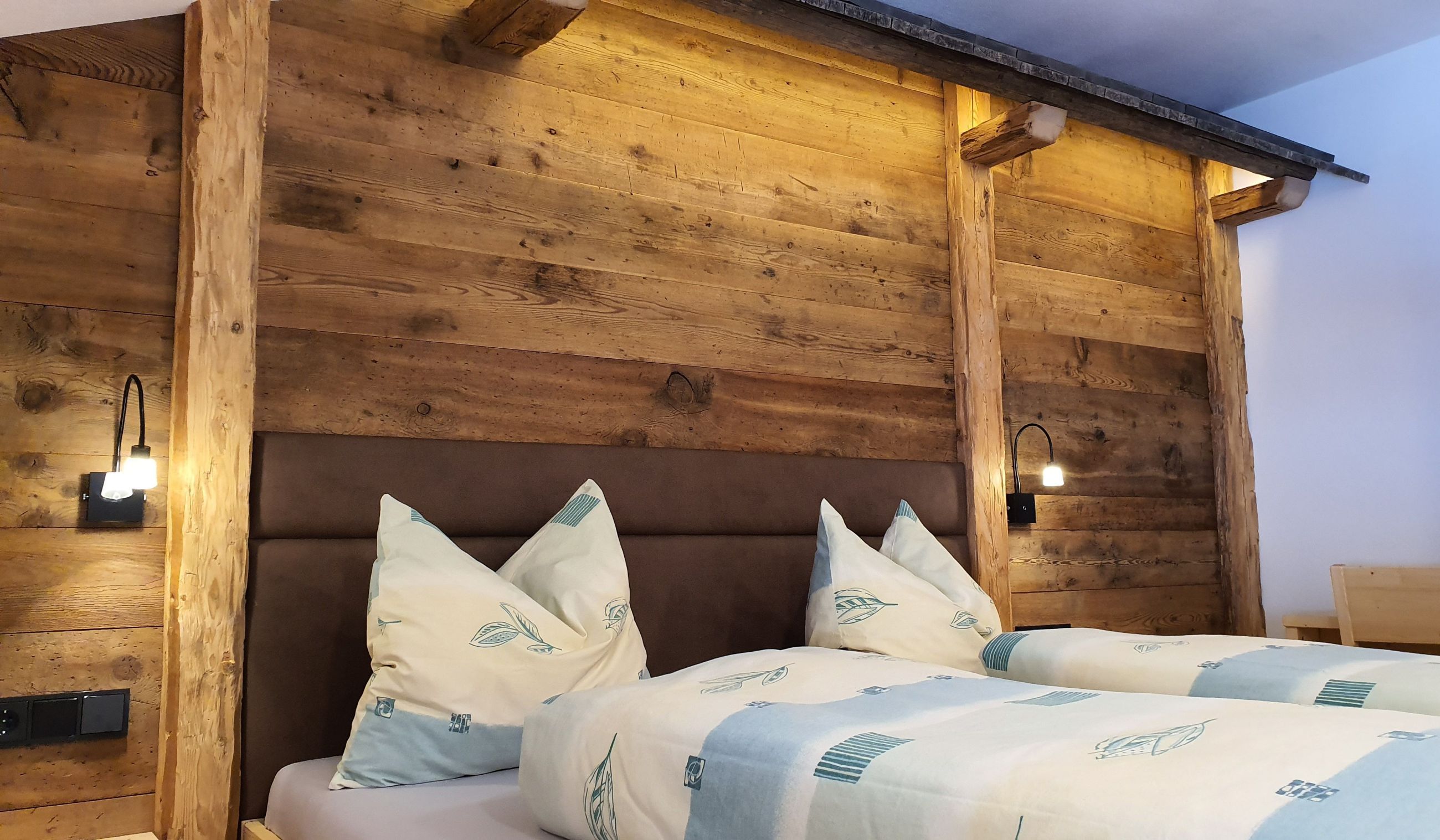 Cabecera de madera con lámpara integrada, cama con ropa de cama blanca y cojines decorativos, y una lámpara fijada a la pared.