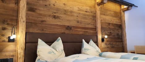 Cabecera de madera con lámpara integrada, cama con ropa de cama blanca y cojines decorativos, y una lámpara fijada a la pared.