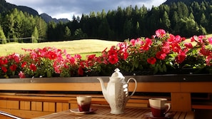 Una mesa de madera con un hervidor blanco y dos tazas, frente a un paisaje montañoso y flores rosadas vibrantes.