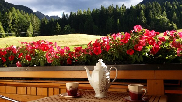 A wooden table with a white teapot and two cups, set against a backdrop of a scenic mountain view and vibrant pink flowers.