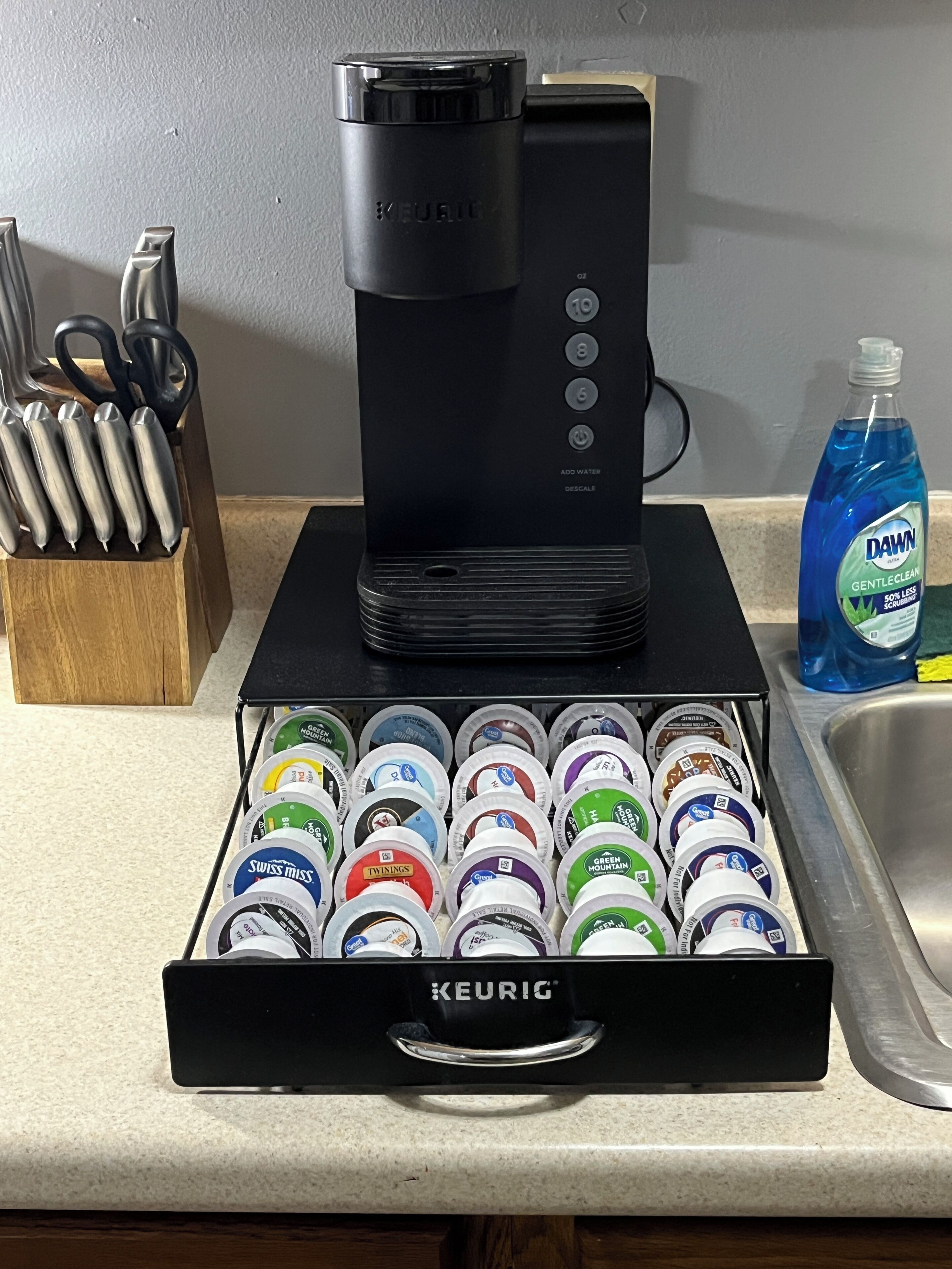 Une machine à café avec un réservoir d’eau posée sur un plan de travail, un tiroir contenant des dosettes de café et une bouteille de liquide vaisselle Dawn.