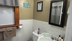 A bathroom with a toilet, sink, and shower. There is a towel rack, a framed sign with bathroom rules, and a window with blinds. - Just Like Being At Home- Newly Renovated Unit (North Troy)