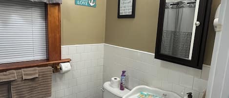 A bathroom with a toilet, sink, and shower. There is a towel rack, a framed sign with bathroom rules, and a window with blinds.