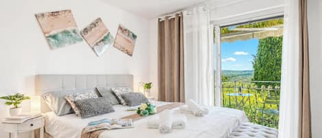 Une chambre à coucher avec un lit, des oreillers, une table de chevet avec une lampe, une fenêtre avec des rideaux et une vue sur la verdure à l’extérieur.